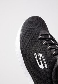 Chaussure de sport noire avec un dessus texturé et des accents blancs, bout rond et semelle en caoutchouc blanc contrastant. Design à lacets.