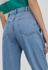 Jean en denim bleu taille haute à coupe ample, avec deux poches arrière, coutures apparentes et texture légèrement délavée.