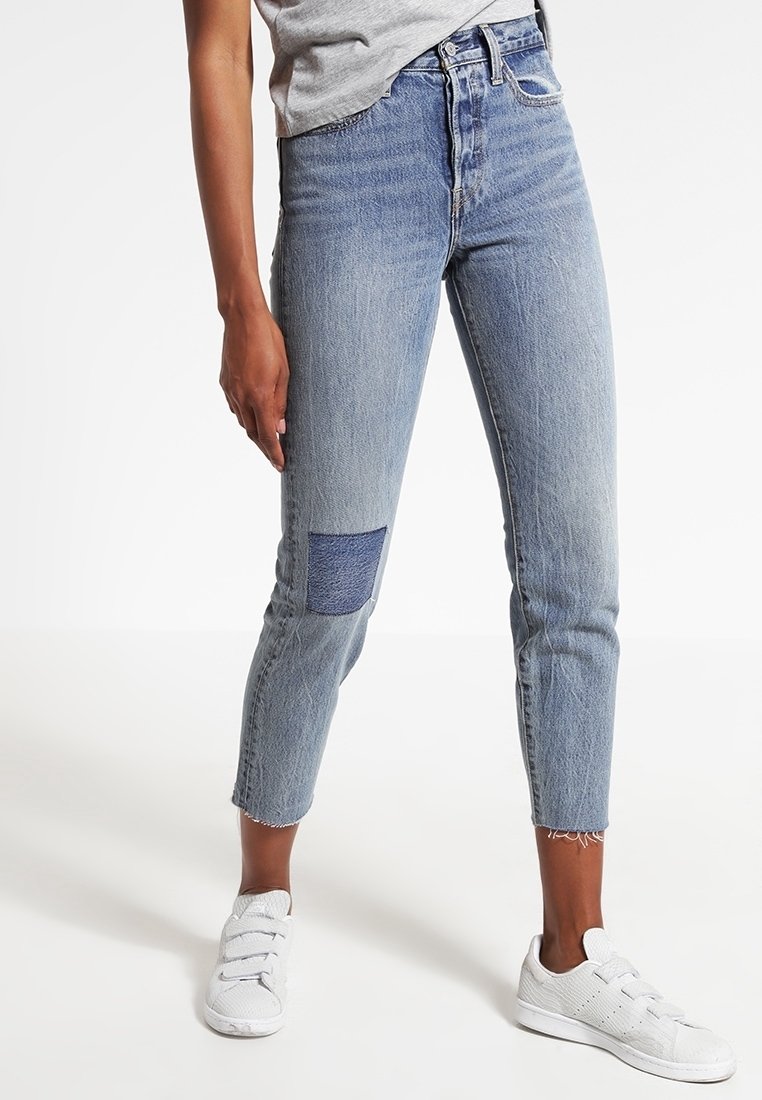 Personne portant un jean court bleu clair avec un patch bleu foncé sur la jambe gauche et des baskets blanches, debout sur un fond blanc.