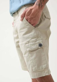 Bermudas cargo beige hechas de tejido ligero, con bolsillos laterales, un detalle de botón y un ajuste relajado. La superficie tiene una textura suave.