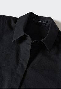 Chemise noire à manches longues avec un col classique pointu, partiellement boutonnée, posée à plat sur une surface claire.