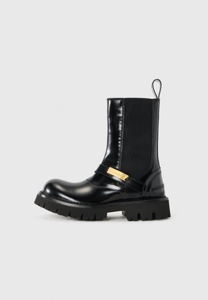 Stivale alla caviglia in pelle nera con pannelli laterali elastici, una fascia con accentuazione dorata e una suola in gomma nera chunky. Finitura liscia con punta rotonda.