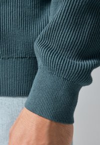 Pull en maille côtelée teal, avec des poignets ajustés et une texture douce. Gros plan sur la manche mettant en avant les détails de construction du vêtement.
