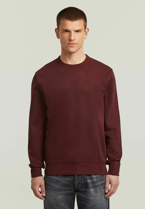 Giovane uomo con capelli corti che indossa una felpa bordeaux semplice e jeans grigi, in piedi davanti a uno sfondo grigio chiaro.