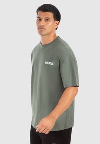 Camiseta de algodón verde con un corte relajado, cuello redondo, mangas cortas y estampado blanco de "SMILODOX" en el lado izquierdo del pecho.