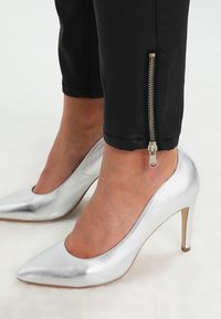 Chaussure à talon haut argentée avec un design à bout pointu et une finition métallique texturée ; portée avec un pantalon noir présentant un détail zippé au niveau de la cheville.