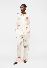 Kobieta stojąca, ubrana w białą koszulę zapinaną na guziki oraz szerokie spodnie z nadrukiem kolorowych koni, połączone z brązowymi butami.