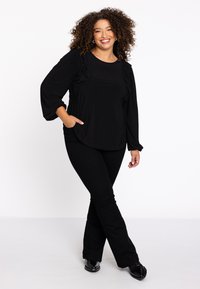 Schwarzes Longsleeve-Oberteil mit rüschierten Akzenten, kombiniert mit schwarzen Schlaghosen. Die Schuhe sind schwarz und glänzend, was einen abgestimmten Look schafft.
