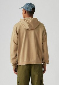 Beige hoodie met een relaxte pasvorm en een grote capuchon, voorzien van ribgebreide manchetten. Gecombineerd met olijfgroene cargobroek. Eenvoudig ontwerp, geen zichtbare patronen.