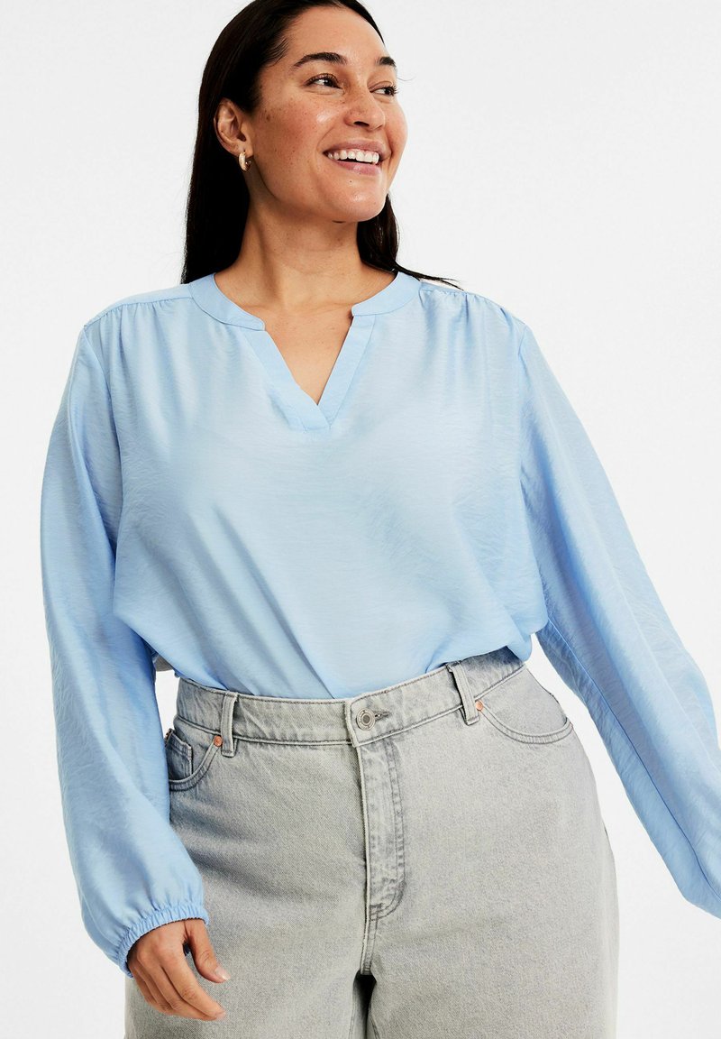 Helle blaue Bluse mit langen Ärmeln und V-Ausschnitt sowie gerafften Schultern, kombiniert mit hochgeschnittenen, hellgrauen Denim-Jeans.