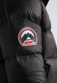 Manche de veste matelassée noire avec un patch rond blanc, rouge et bleu représentant un graphisme de montagne et le texte "JACKIT" en rouge. Tissu lisse et brillant.