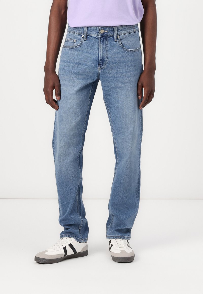 Jean en denim bleu clair avec une coupe droite, poches avant et une finition légèrement délavée, portés avec des baskets blanches et grises.