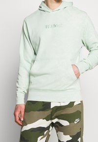 Persona che indossa un felpa verde chiaro con la scritta "JUST DO IT." e pantaloni Nike con motivo mimetico, con una mano in tasca della felpa.