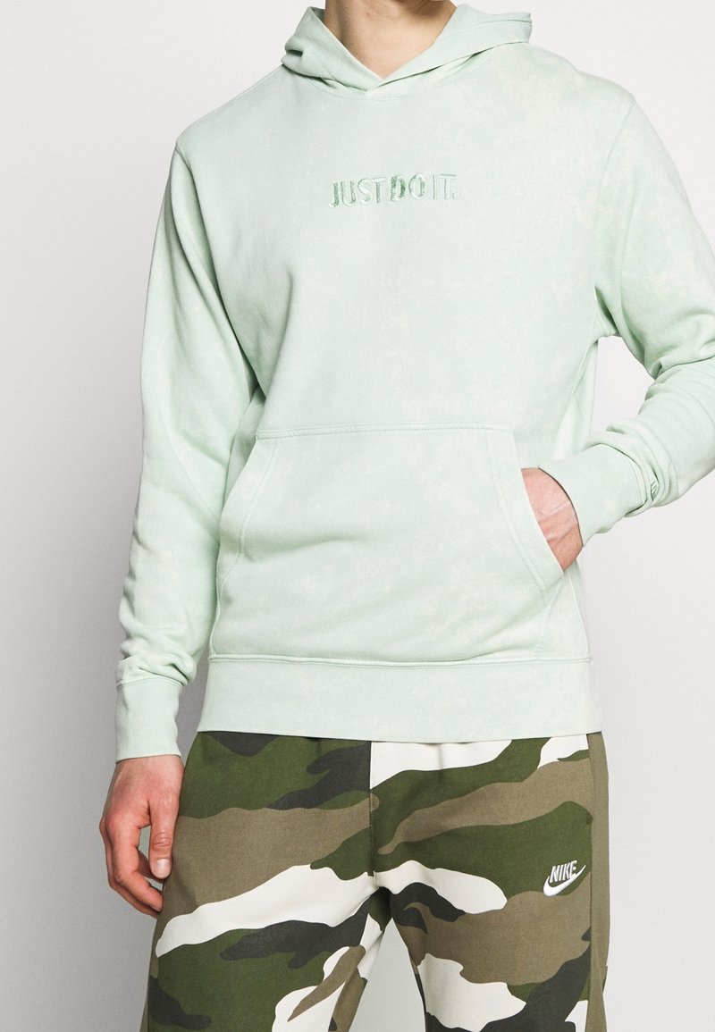Persona che indossa un felpa verde chiaro con la scritta "JUST DO IT." e pantaloni Nike con motivo mimetico, con una mano in tasca della felpa.