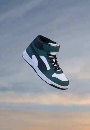 Basket montante avec des panneaux verts, noirs et blancs, des lacets noirs et une sangle Velcro sur un fond de ciel nuageux.