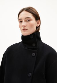 Schwarzer Wollmantel mit hohem Kragen, der vorne fünf große Knöpfe hat. Der Stoff hat eine glatte Textur und ein minimalistisches Design.