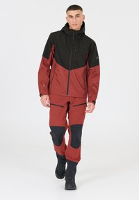 Röd och svart vattentät jacka med huva, utrustad med en ficka med dragkedja på bröstet och ytterligare sidofickor med dragkedja; designad för utomhusaktiviteter.