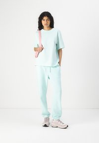 Mintgroene oversized t-shirt en bijpassende joggingbroek, met een comfortabele pasvorm. Model houdt een lichtroze schoudertas vast en draagt lichte grijze sneakers.