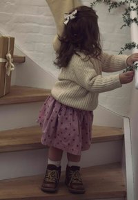 Barn iført en cremefarvet strikket sweater og en lyserød nederdel med prikker, kombineret med brune støvler og hvide sokker, der går op ad trætrappen.