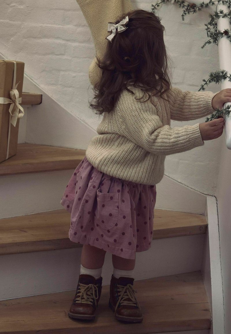 Barn iført en cremefarvet strikket sweater og en lyserød nederdel med prikker, kombineret med brune støvler og hvide sokker, der går op ad trætrappen.
