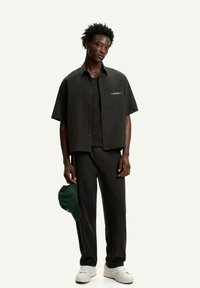Camicia nera a maniche corte con bottoni e taschino anteriore, abbinata a pantaloni neri e sneakers bianche; tiene in mano un cappellino verde.