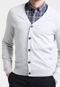 Ljusgrå cardigan med V-ringning, svarta knappar, ribbade ärmslut och nederkant, bärs över en marinblå rutig skjorta med vita detaljer.