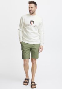 Felpa crema con logo ricamato, abbinata a pantaloni corti verdi e sandali marroni. Outfit casual caratterizzato da una vestibilità comoda e dettagli minimi.