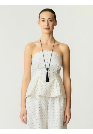 Kvinde står iført hvid stropløs peplum-top og bukser med små sorte prikker, sort halskæde med kvast, neutral baggrund.