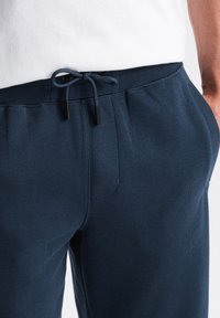 Námořnické modré tepláky s elastickým pasem, nastavitelnou šňůrkou, bočními kapsami a hladkým textilním povrchem. Viditelné zvýšené švy.