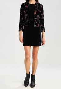 Chaqueta cropped de terciopelo negro con bordado floral, combinada con un vestido negro sin mangas y botas hasta el tobillo. Textura suave y diseño ajustado.