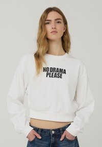 Giovane con lunghi capelli biondi che indossa un crop top bianco con la scritta "NO DRAMA PLEASE" e jeans blu, mani in tasca.