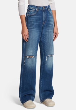 Personne portant un jean bleu large avec des déchirures aux genoux, un haut blanc à manches longues et des chaussures beige à enfiler, debout devant un fond uni.