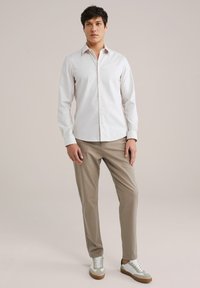 Camisa blanca de manga larga con botones combinada con chinos beige y zapatillas blancas con detalles en gris y marrón, sobre un fondo neutro.