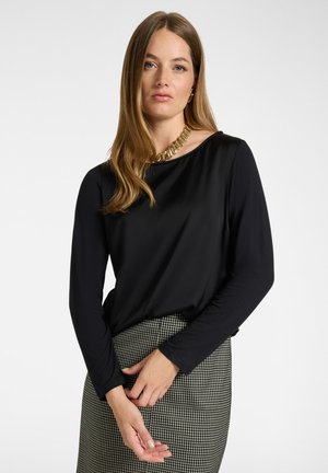 Vrouw met lang lichtbruin haar, die een zwarte top met lange mouwen, een gouden ketting en een zwart-witte geruite rok draagt, staand met gevouwen handen.