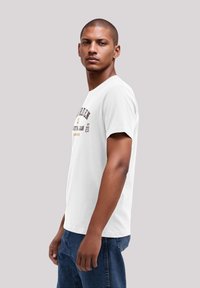 Weißes Baumwoll-T-Shirt mit einem bedruckten Design in dunklem und goldenem Text, Rundhalsausschnitt und kurzen Ärmeln, kombiniert mit dunkelblauen Jeans.