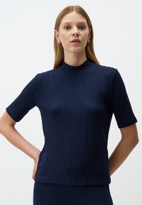 Marineblauw T-shirt met korte mouwen, hoge hals, geribbelde textuur en subtiel geometrisch patroon; aansluitende snit en zachte stof.