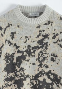 Pull en tricot gris clair avec un motif tacheté noir et beige. Il possède un col côtelé et une texture douce. Étiquette de la marque visible.
