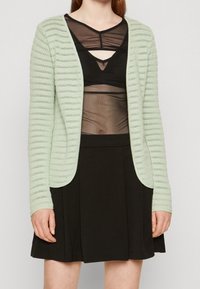 Mátově zelený pletený cardigan s texturovanými pruhy, otevřeným předním designem a dlouhými rukávy, spárovaný s průhledným černým topem a černou sukní.