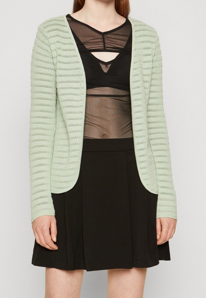 Mátově zelený pletený cardigan s texturovanými pruhy, otevřeným předním designem a dlouhými rukávy, spárovaný s průhledným černým topem a černou sukní.