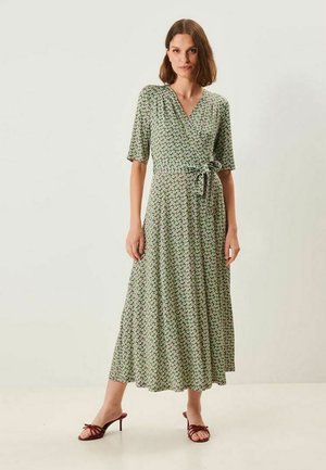 Kvinde med skulderlangt brunt hår iført en grøn blomsterprint slå-om-kjole med albuelange ærmer og røde åbne hælesandaler.