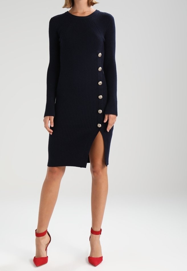 Marineblaues geripptes Strickkleid mit langen Ärmeln, seitlichen Knopfdetails, hohem Schlitz und rundem Ausschnitt. Kombiniert mit roten High-Heels.