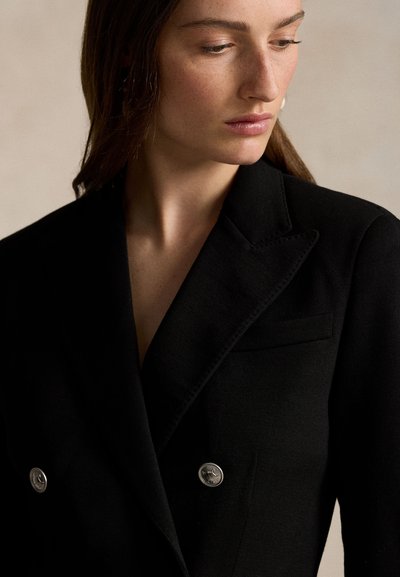 O femeie îmbrăcată într-un sacou negru cu două rânduri de nasturi argintii, privind în jos și spre lateral, pe un fundal bej simplu.