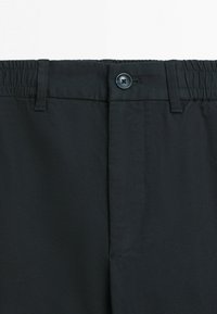 Zbliżenie na pas czarnych spodni z elastycznymi bokami, zapięciem na guzik i szlufkami na pasek na białym tle.