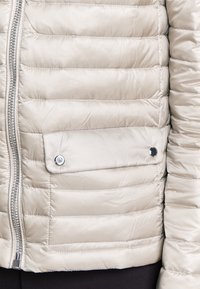 Light beige padded jacket with horizontal quilted design, silver zipper, and a snap button pocket on the front. Smooth, shiny texture.
