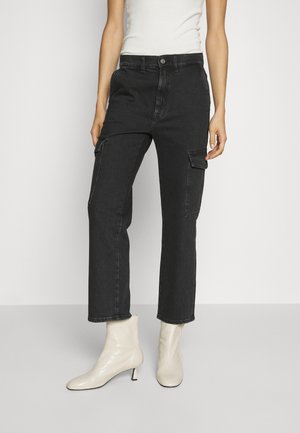 Vaqueros cargo negros de corte slim con bolsillos con solapa, combinados con botines blancos y una camiseta sin mangas blanca, de pie frente a un fondo liso.