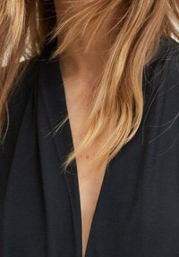 Abbigliamento nero a collo a V con una finitura testurizzata. Capelli ondulati e sciolti parzialmente visibili, creando un aspetto morbido attorno al collo.
