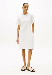 Robe en coton blanc à manches courtes, silhouette droite et poches latérales. Associée à des sandales crème avec une boucle décorative.