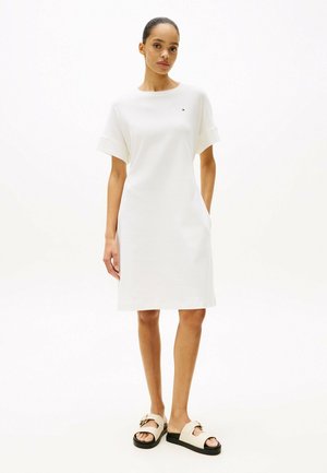 Robe en coton blanc à manches courtes, silhouette droite et poches latérales. Associée à des sandales crème avec une boucle décorative.