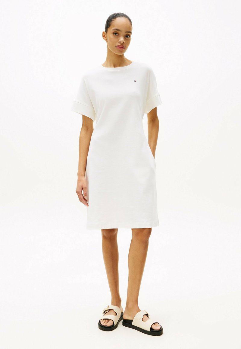 Robe en coton blanc à manches courtes, silhouette droite et poches latérales. Associée à des sandales crème avec une boucle décorative.