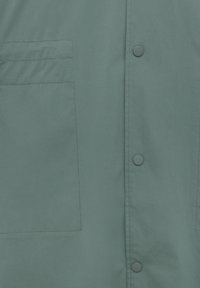 Tissu gris-vert avec une grande poche rectangulaire, un petit détail cousu horizontalement et trois boutons-pression ronds sur le côté droit.